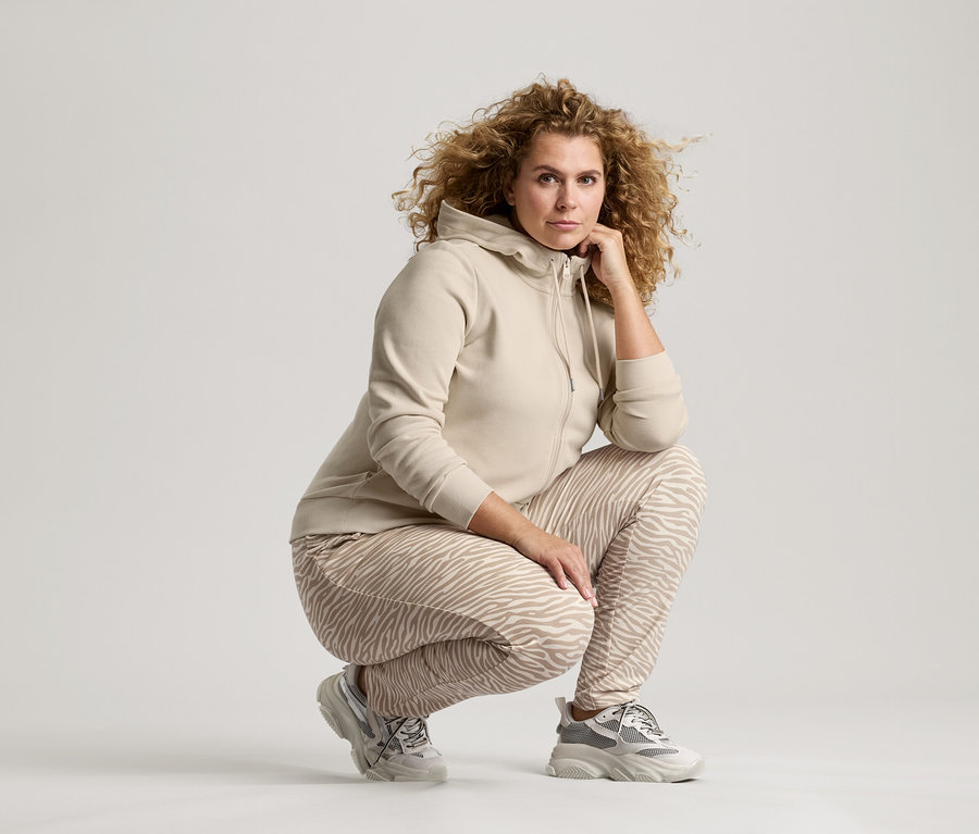 Žena s kudrnatými vlasy v béžové bundě, kalhotách se vzorem zebry a teniskách se krčí s rukou na bradě.
