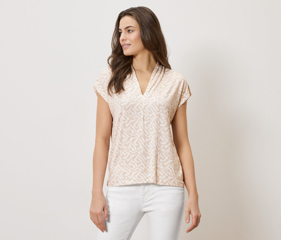 Žena má na sobě světle hnědou vzorovanou halenku s výstřihem do V a bílé kalhoty.