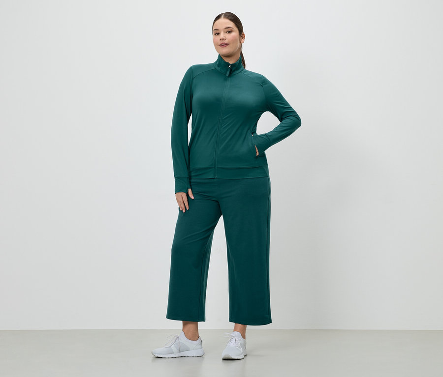 Žena ve smaragdově zelené teplákové soupravě s bundou na zip a culottes kalhotami a bílých teniskách.