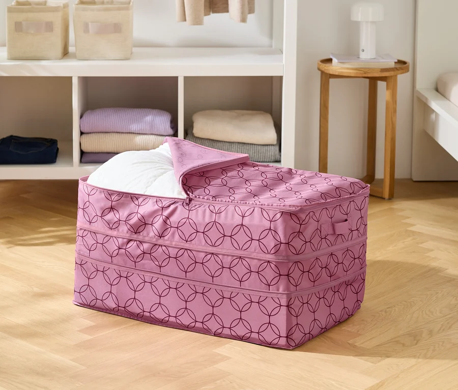 Na dřevěné podlaze stojí růžová úložná taška s geometrickým vzorem, částečně vyplněná bílou dekou.