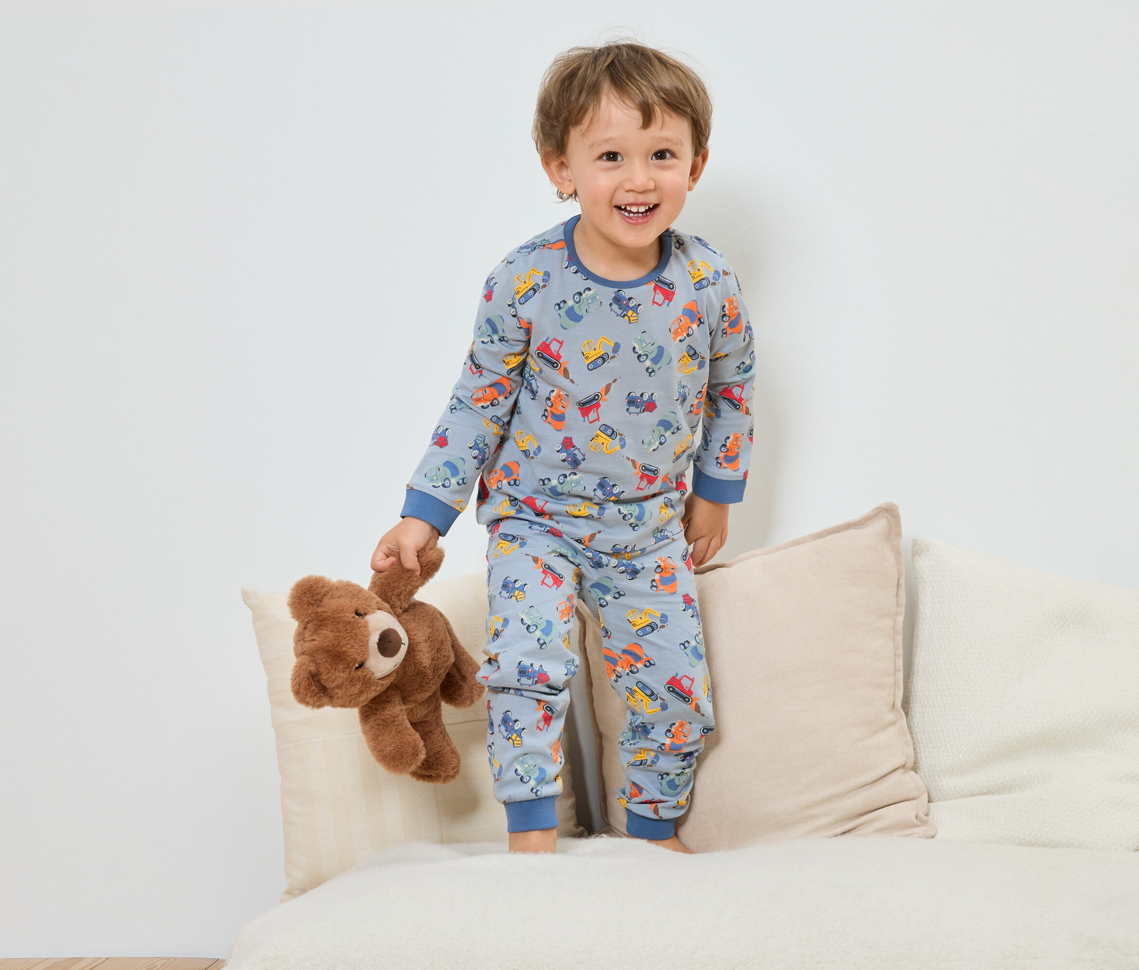 Usmívající se chlapec v modrém pyžamu se vzorem stavebních vozidel stojí s plyšovým medvídkem na pohovce.