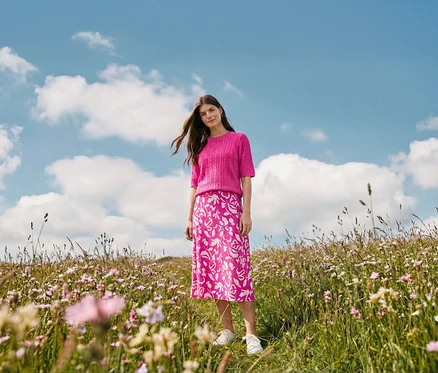 Žena stojí na louce a má na sobě midi sukni s potiskem, náušnice Wave a kožené tenisky.