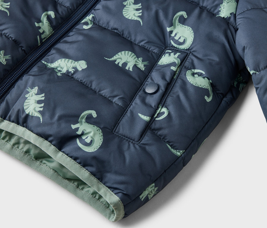 Detailní záběr na tmavě modrou prošívanou bundu se světle zeleným vzorem dinosaurů, zipem a kapsou na patentku.