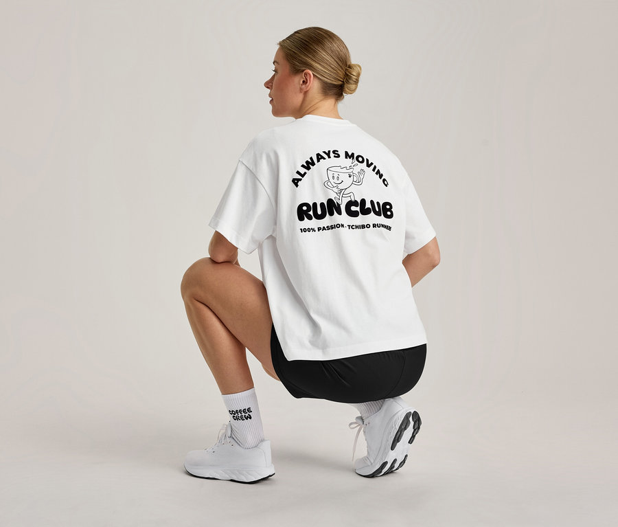 Žena s blond drdolem dřepí, viděná zezadu. Má na sobě bílé, oversized sportovní tričko s černou grafikou 'ALWAYS MOVING RUN CLUB' na zádech, černé šortky, bílé ponožky a bílé sportovní boty.