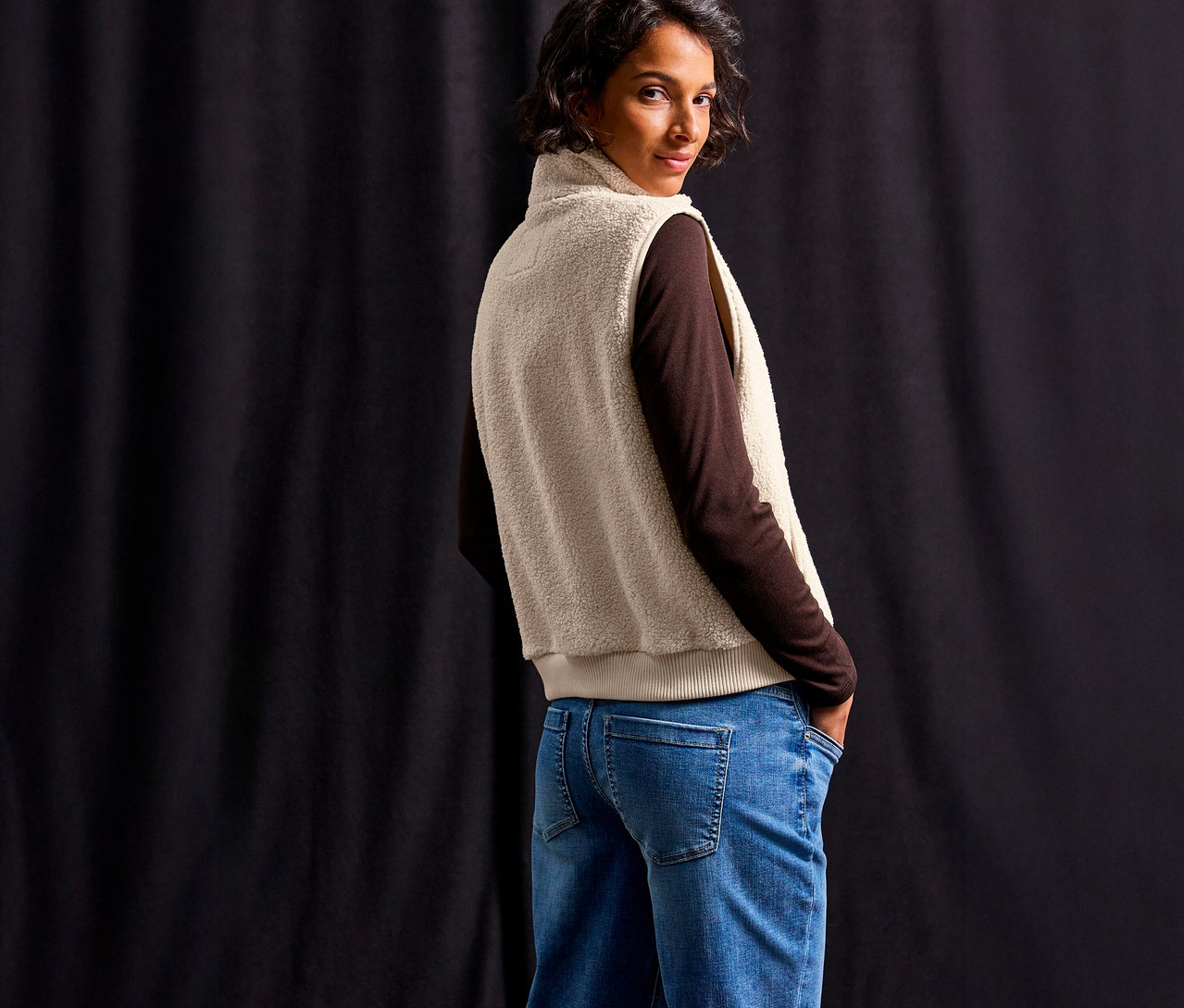 Žena v hnědém topu a modrých džínách má na sobě plyšovou vestu Street One a dívá se přes rameno.