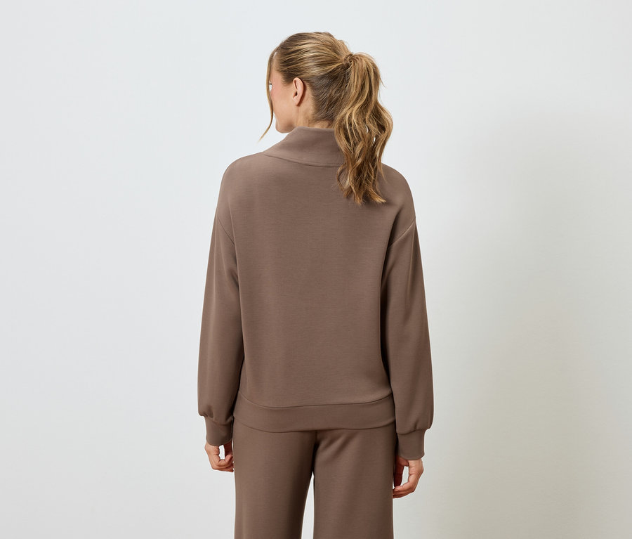 Pohled zezadu na ženu s blond vlasy v hnědém svetru a hnědých kalhotách.