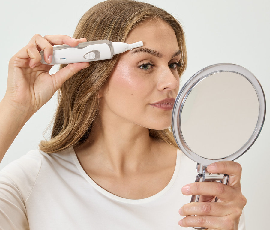 Žena si upravuje obočí bílým epilátorem a dívá se do kulatého zrcadla.