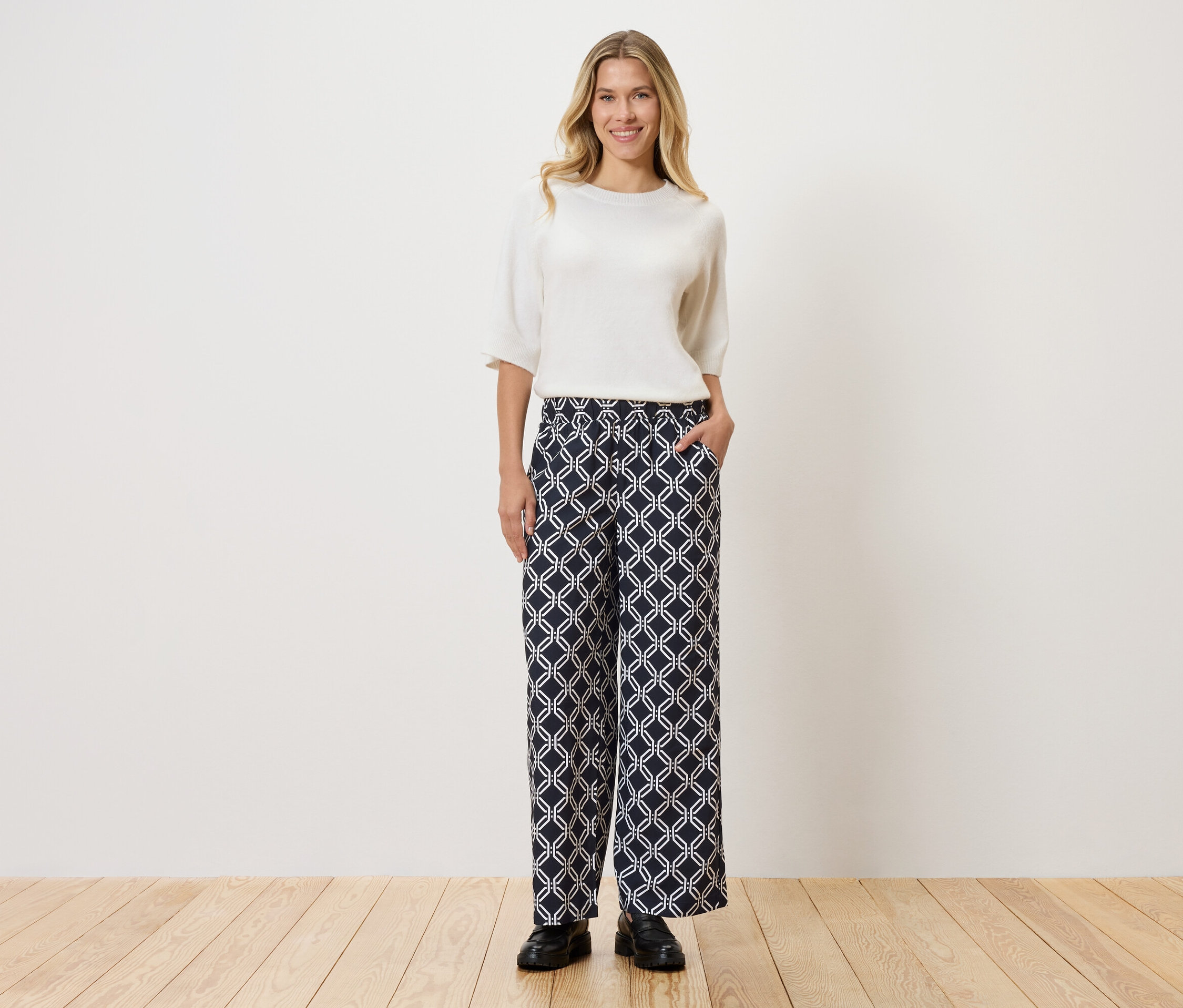 Žena v bílém topu a vzorovaných kalhotách stojící na dřevěné podlaze před zdí.