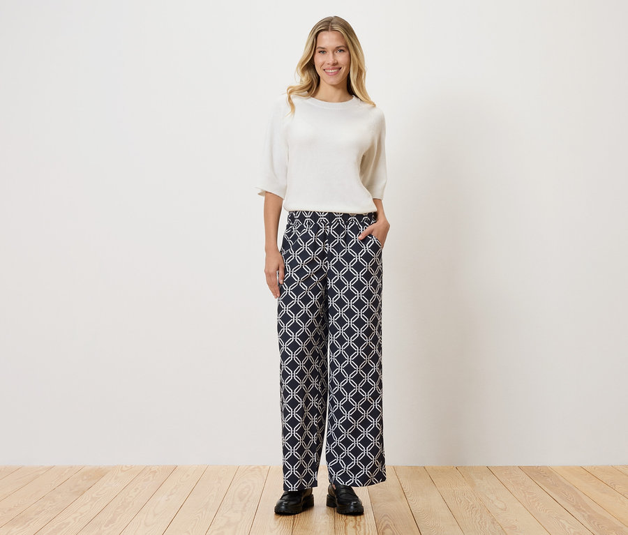 Žena v bílém topu a vzorovaných kalhotách stojící na dřevěné podlaze před zdí.