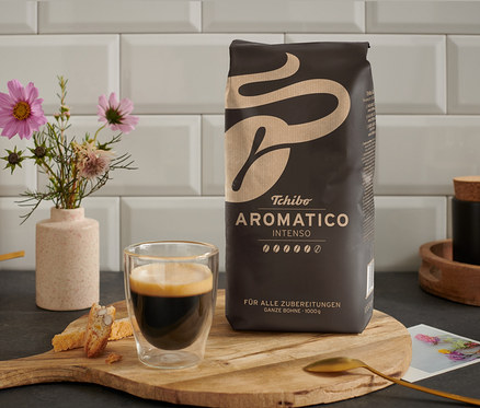 Na dřevěném stole stojí šálek kávy vedle balení Aromatico Intenso - 1 kg zrnkové kávy.