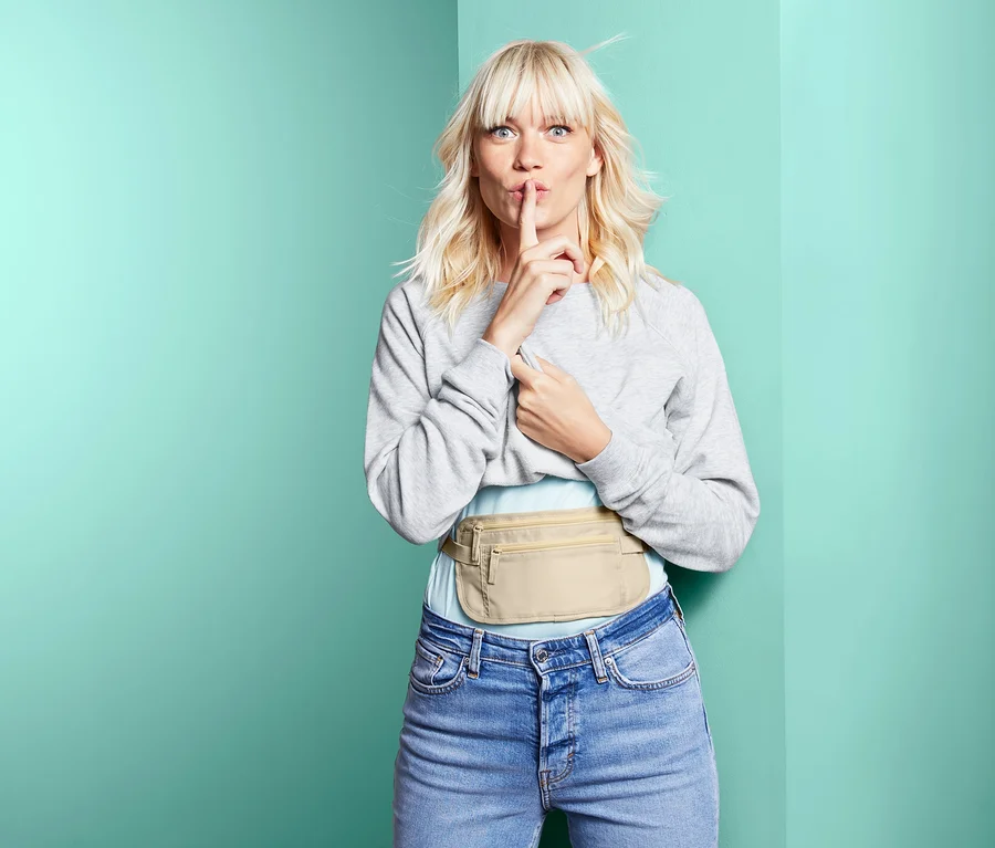 Blonďatá žena dává prst na ústa a nosí béžovou bezpečnostní ledvinku.