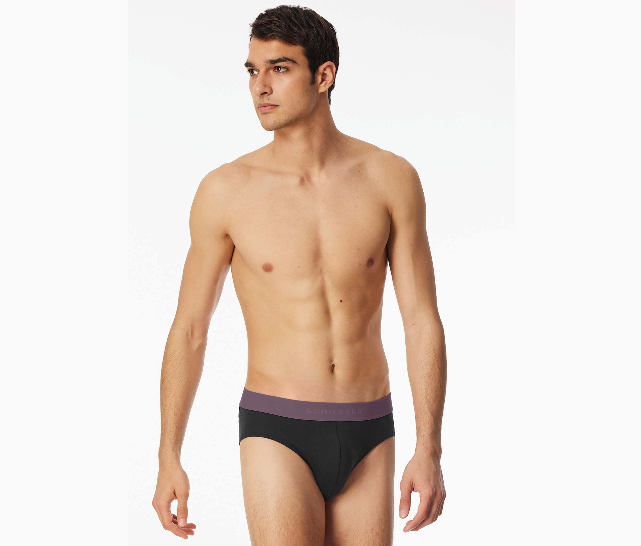 Muž s odhalenou horní částí těla nosí černé slipy Schiesser s fialovým pasem.