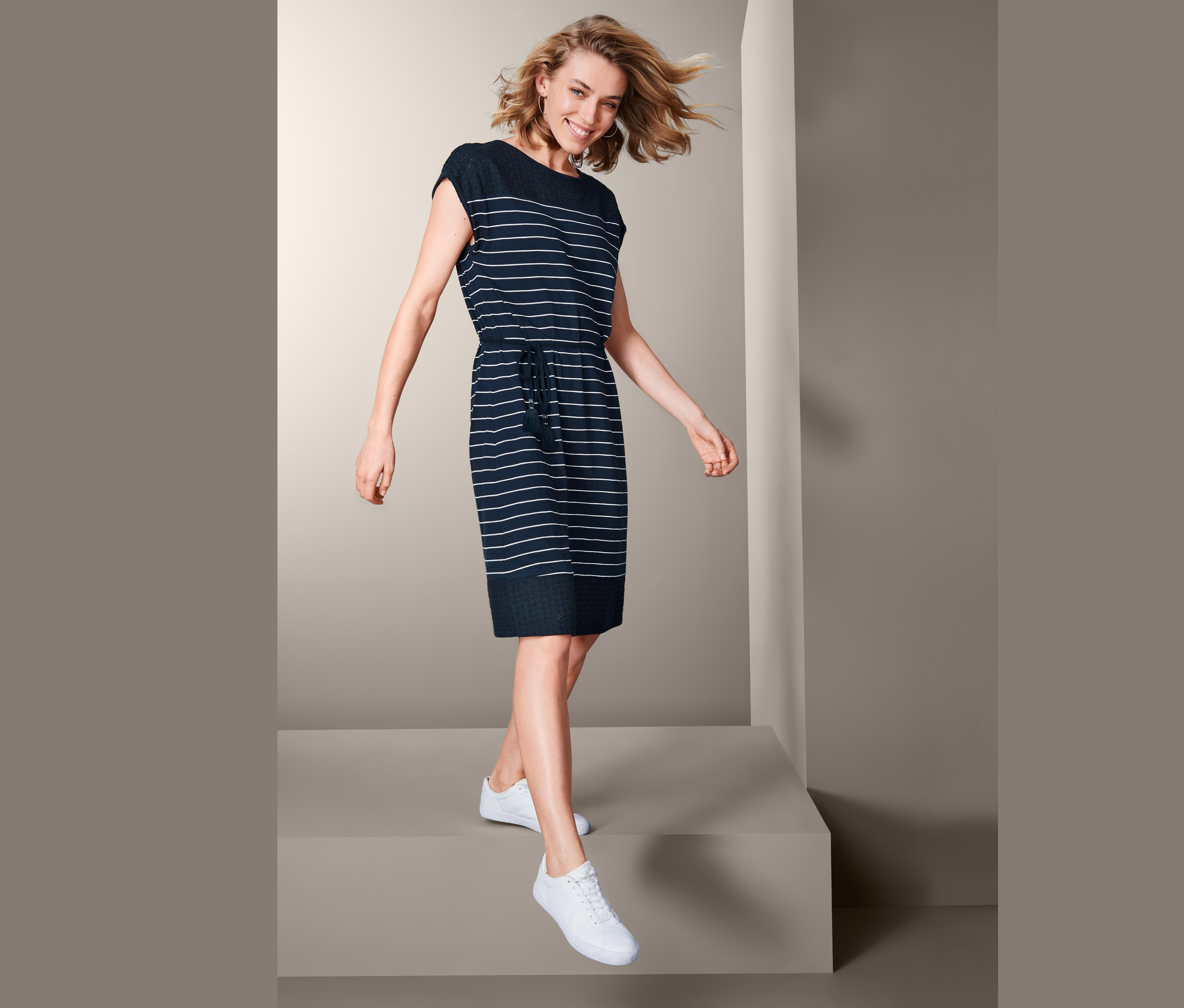 Žena kráčí a má na sobě žerzejové šaty s ažurovou výšivkou a bílé tenisky.