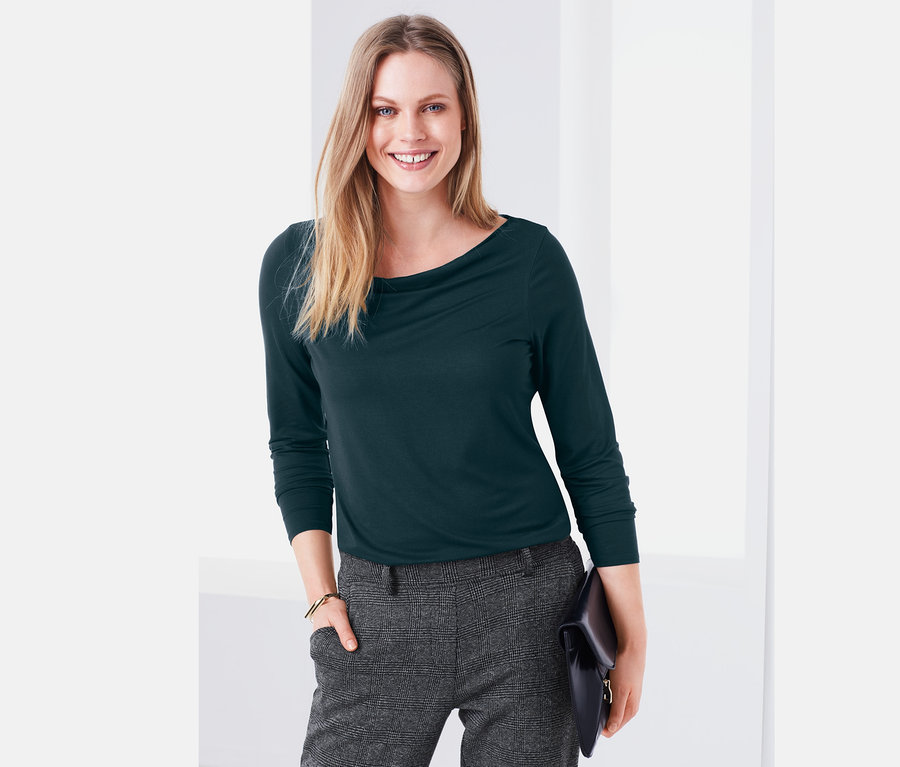 Žena má na sobě zelené tričko s výstřihem „voda“ a šedé kalhoty.