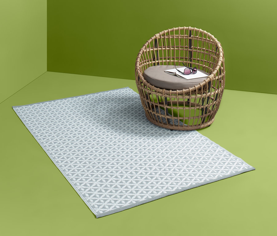 Vzorovaný venkovní koberec leží vedle proutěného křesla v zeleném rohu místnosti.