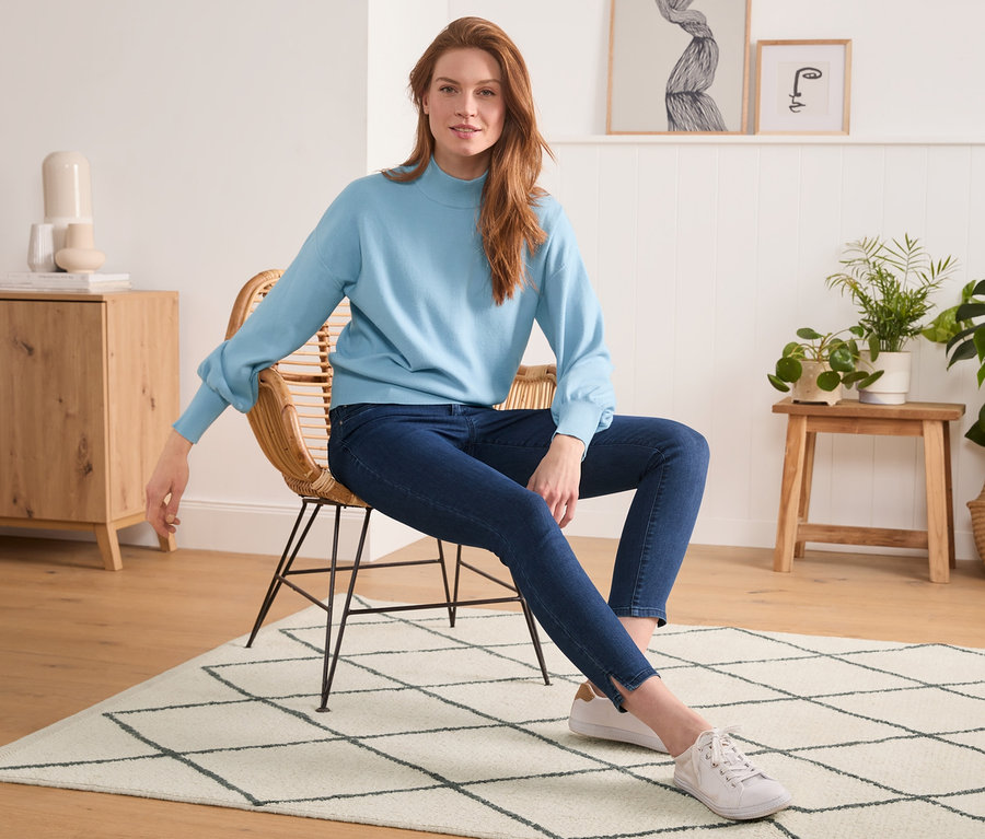 Žena sedí na proutěném křesle na koberci a má na sobě modrý svetr a Skinny džíny.