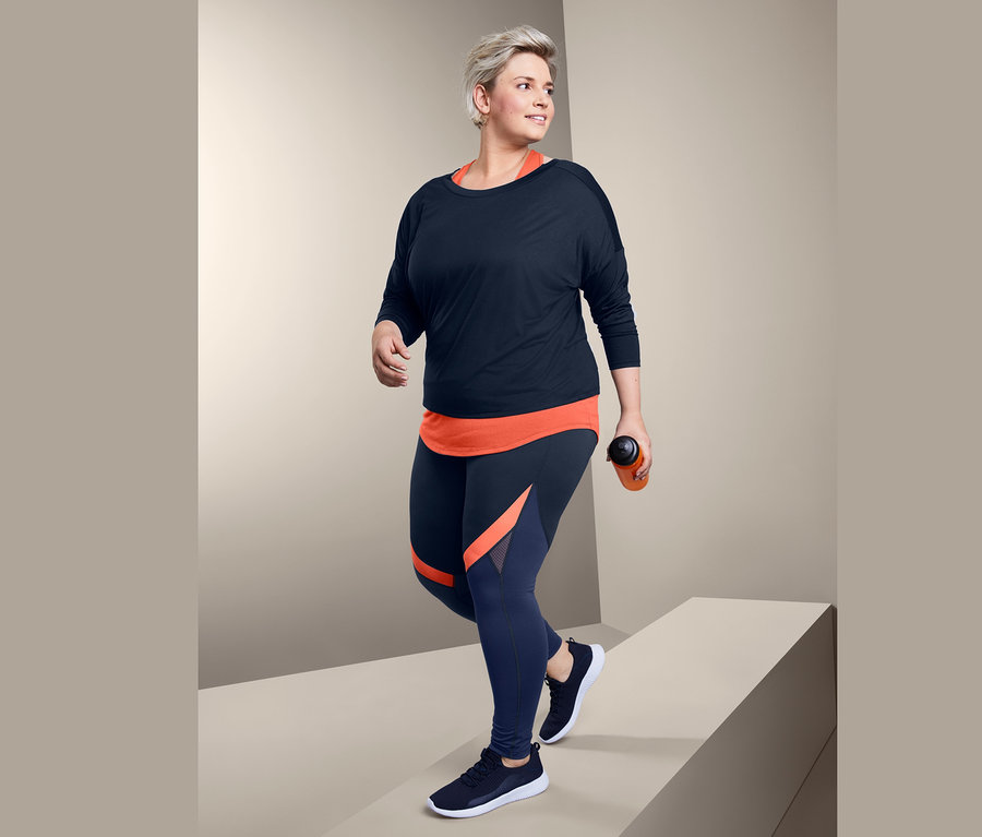 Žena kráčí a má na sobě sportovní triko 2 v 1 a sportovní legíny ve stylu color blocking.