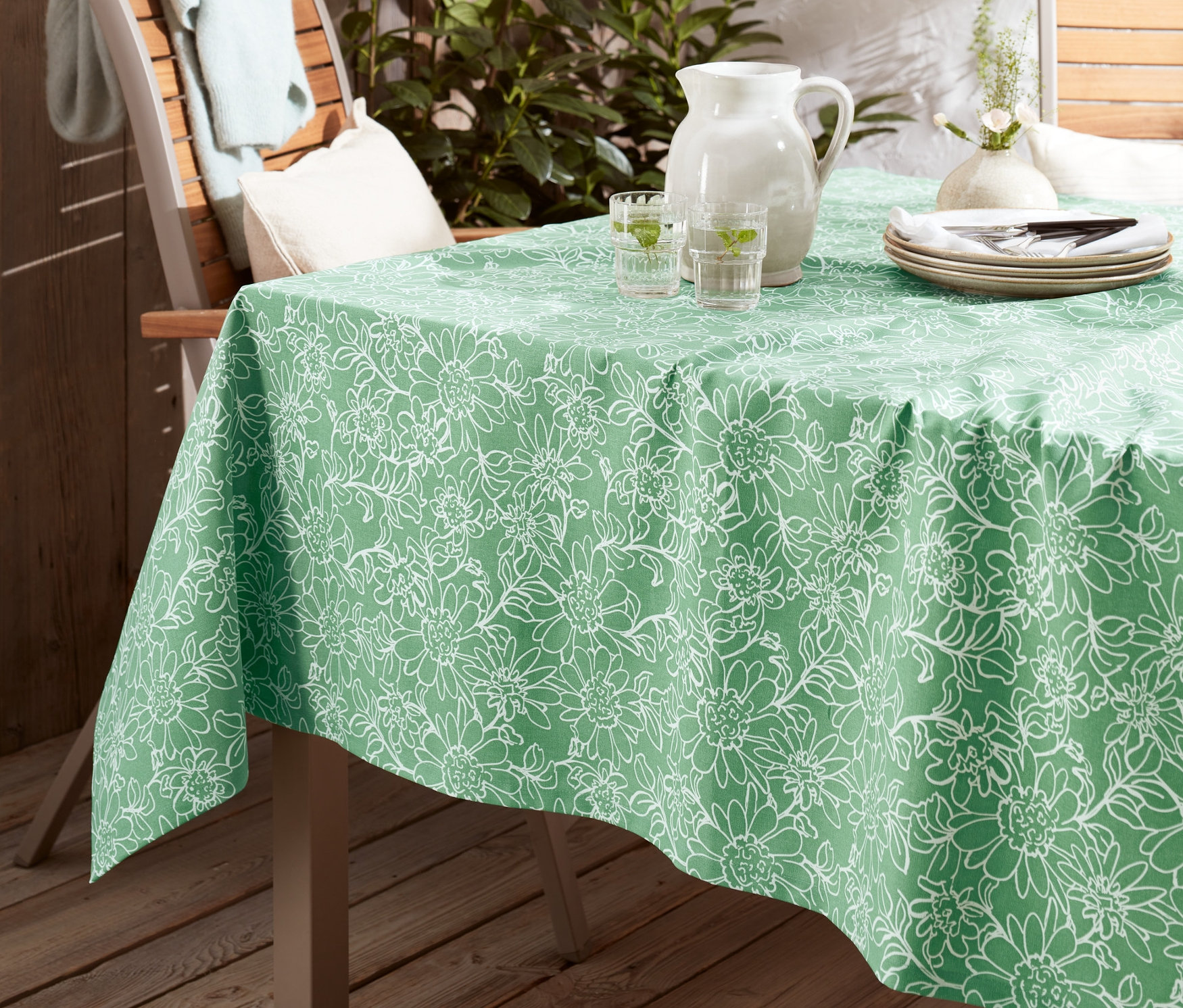 Zelený venkovní ubrus s květinovým vzorem na stole. Na stole stojí džbán s vodou a sklenice.