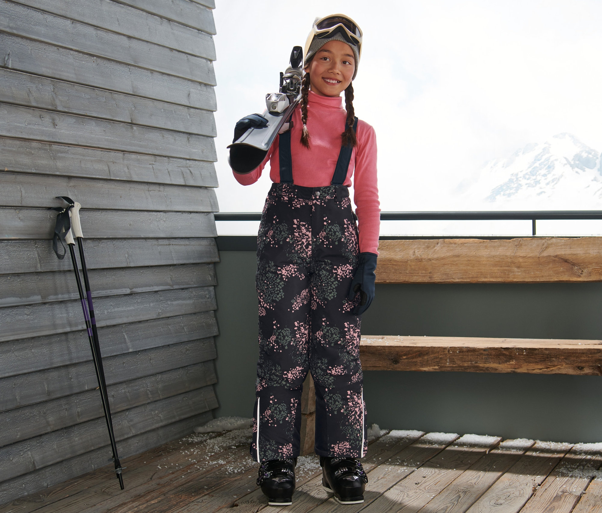 Dívka stojí a nese lyže přes rameno. Má na sobě lyžařské kalhoty s celopotiskem.