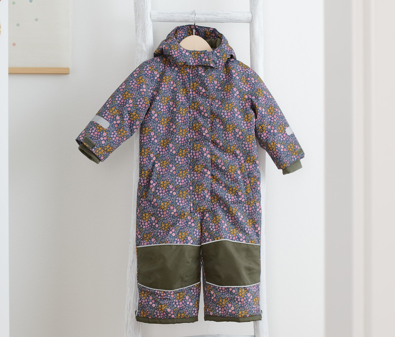 Zimní kombinéza s květovaným potiskem visí na bílém žebříku, vedle jsou žluté boty.