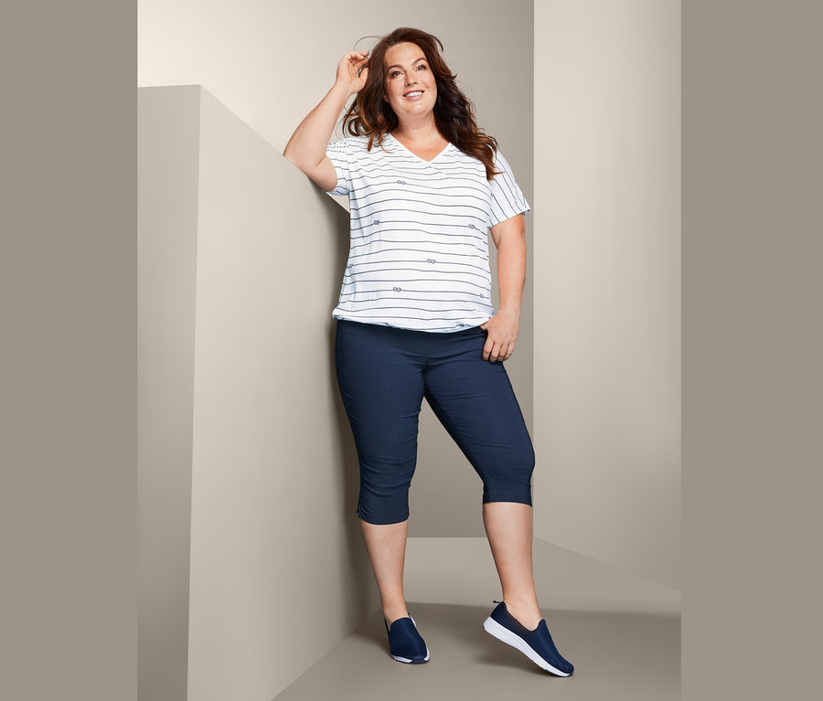 Žena opírající se o zeď má na sobě halenkové triko s tříčtvrtečními bengalinovými kalhotami.