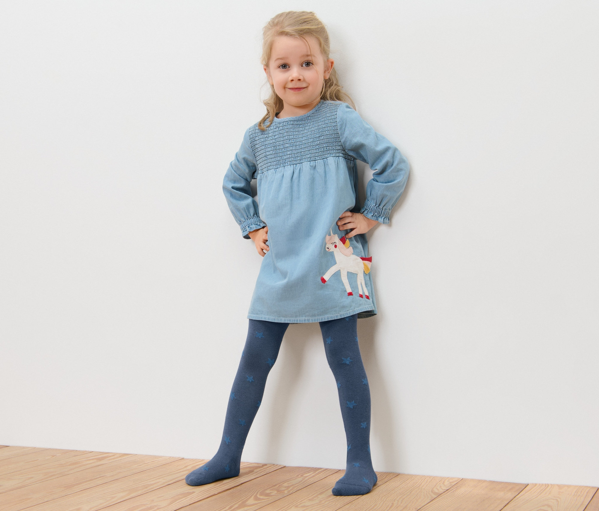 Holčička pózuje v modrých šatech s jednorožcem a bavlněných punčocháčích s hvězdičkami (5 ks).