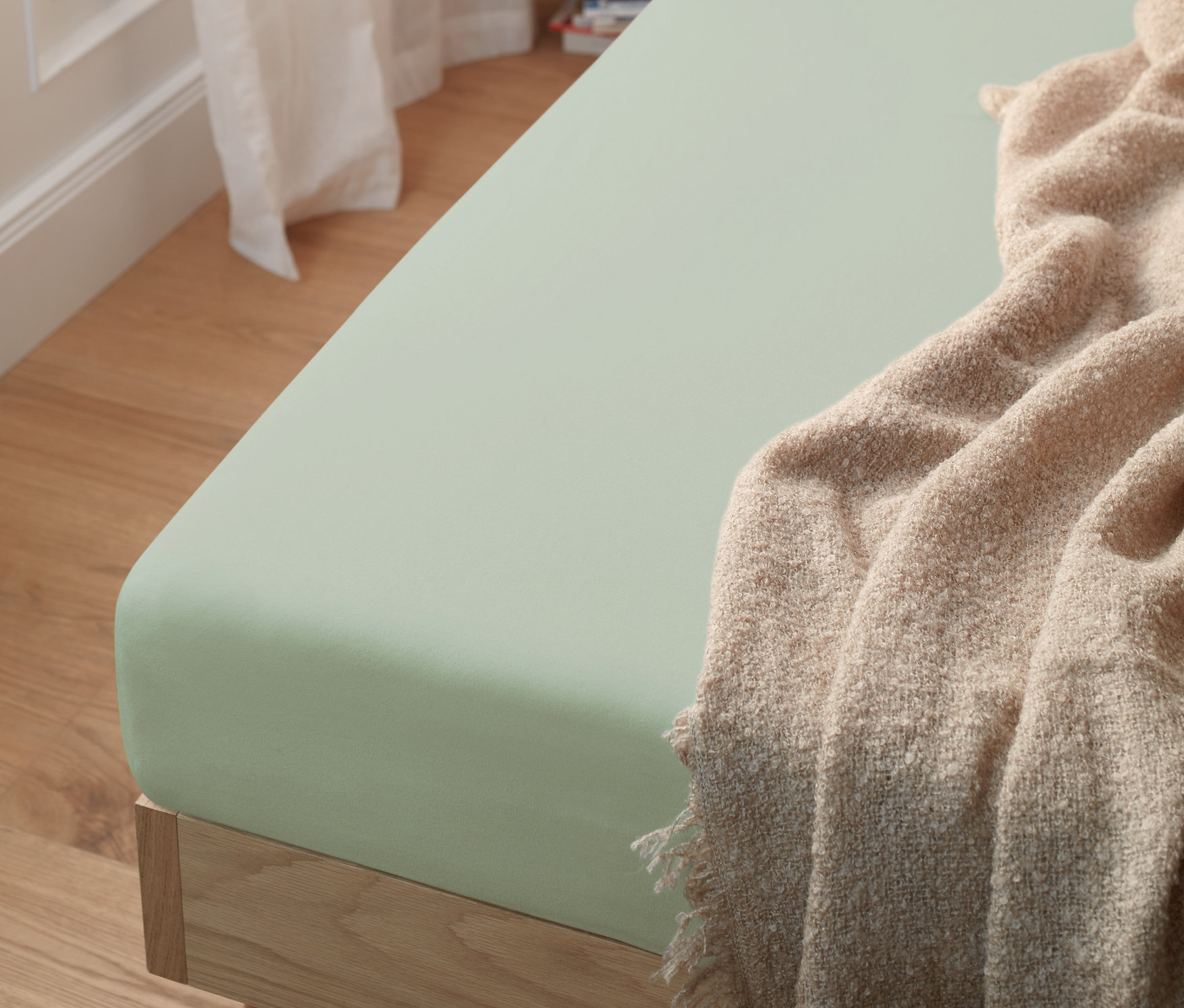 Zelené žerzejové napínací prostěradlo na posteli. Přehoz leží na rohu postele.