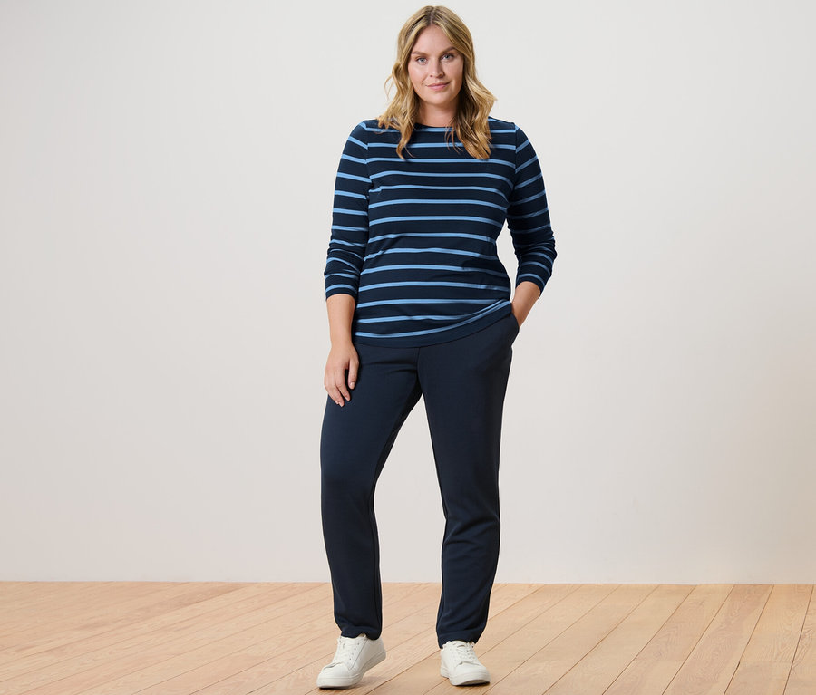 Žena stojí a má na sobě tmavě modré joggpant kalhoty a proužkované triko s dlouhým rukávem.