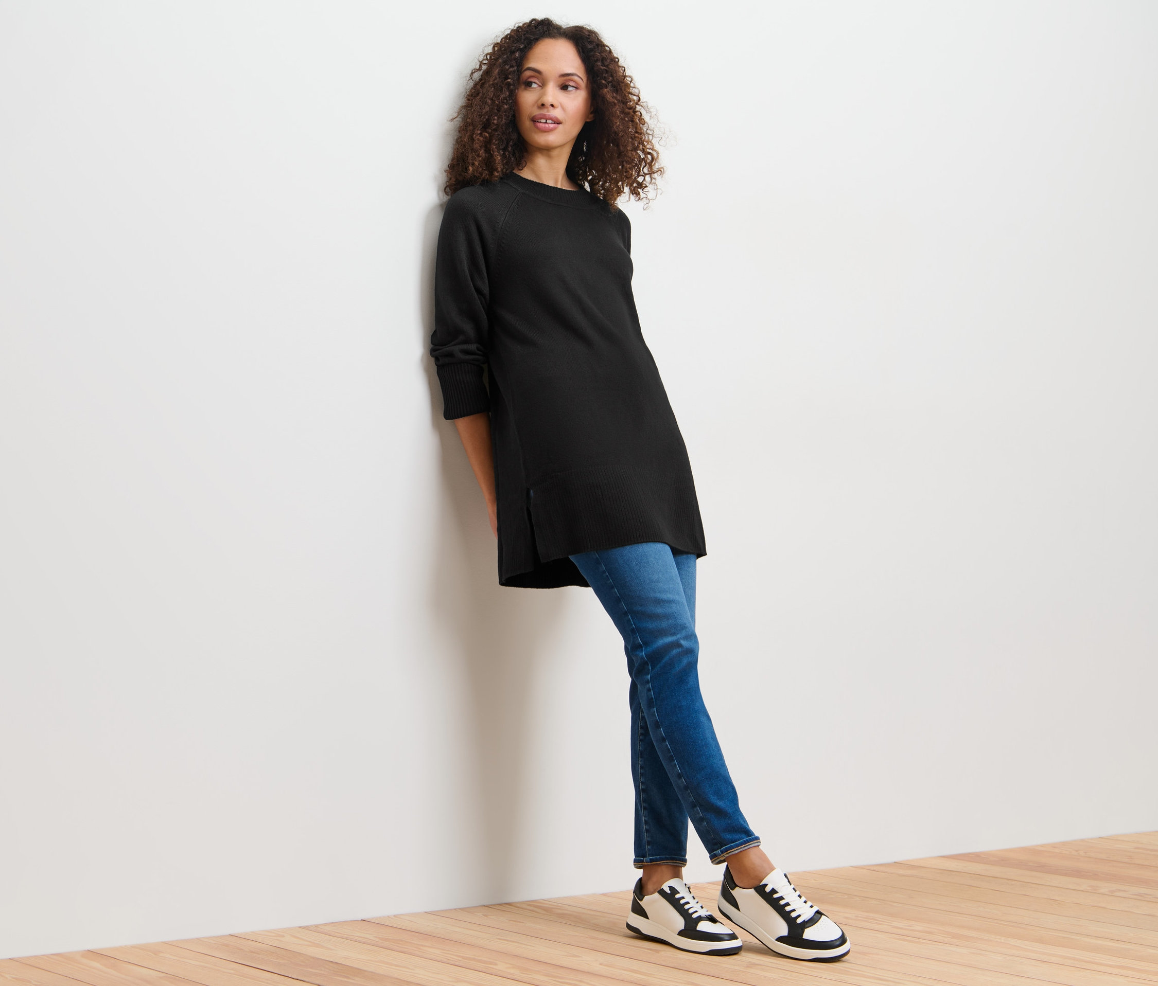 Žena se opírá o stěnu a má na sobě dlouhý černý pletený svetr.