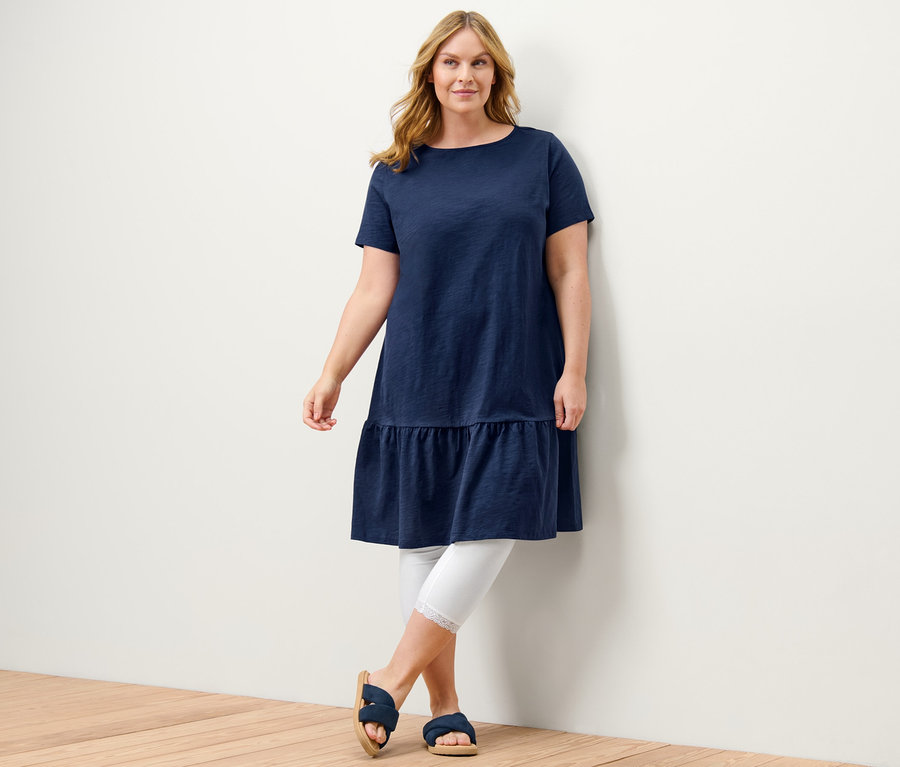 Žena pózuje v tmavě modrých žerzejových šatech s volánem a bílých 3/4 legínách s krajkovým lemem.