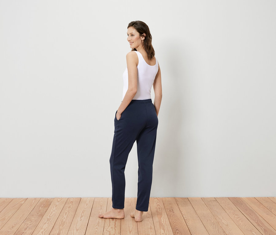 Žena v bílém tílku a tmavě modrých kalhotách stojí zády k divákovi a dívá se přes rameno.