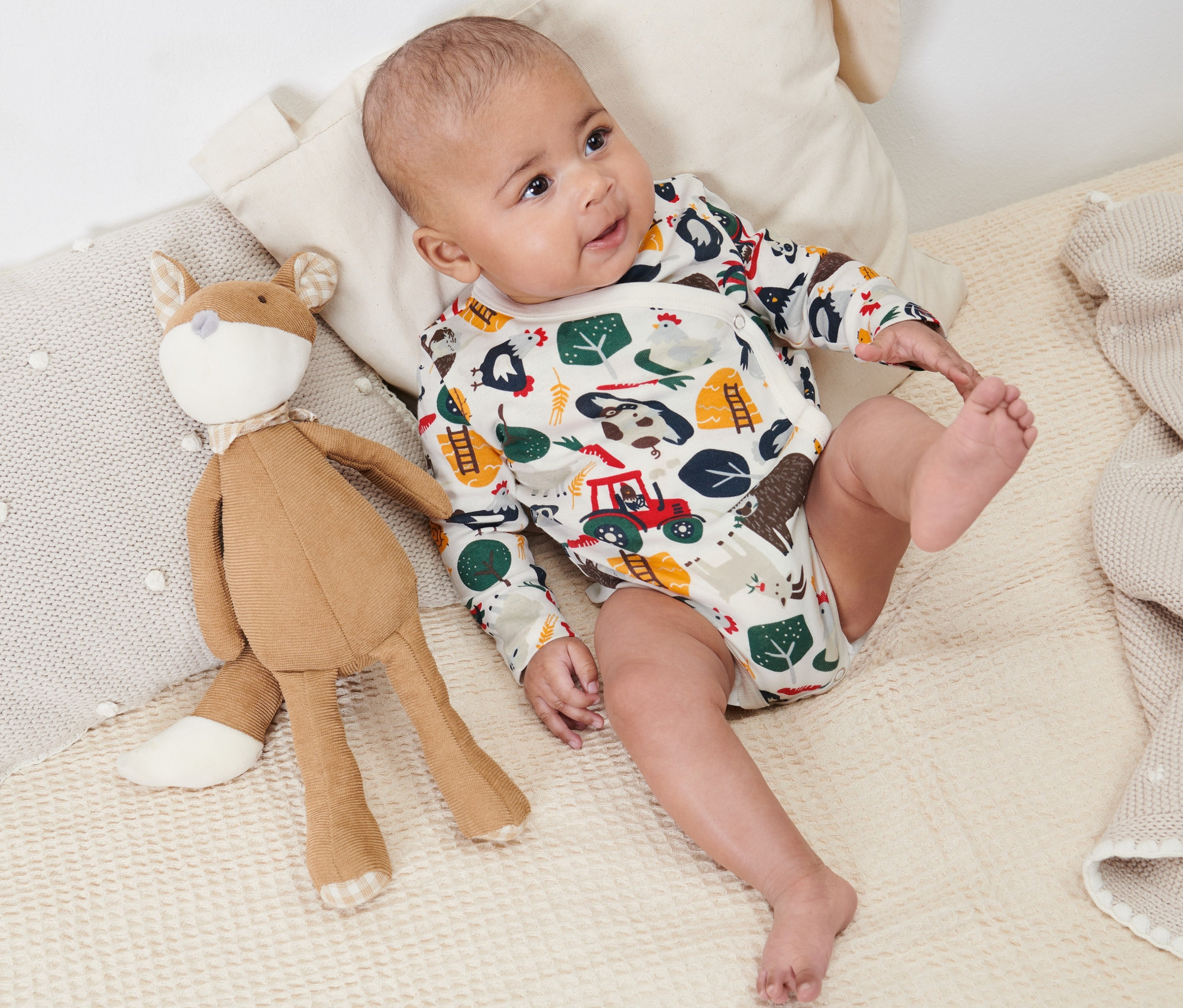 Dítě leží na dece vedle plyšové lišky a má na sobě body s dlouhým rukávem.