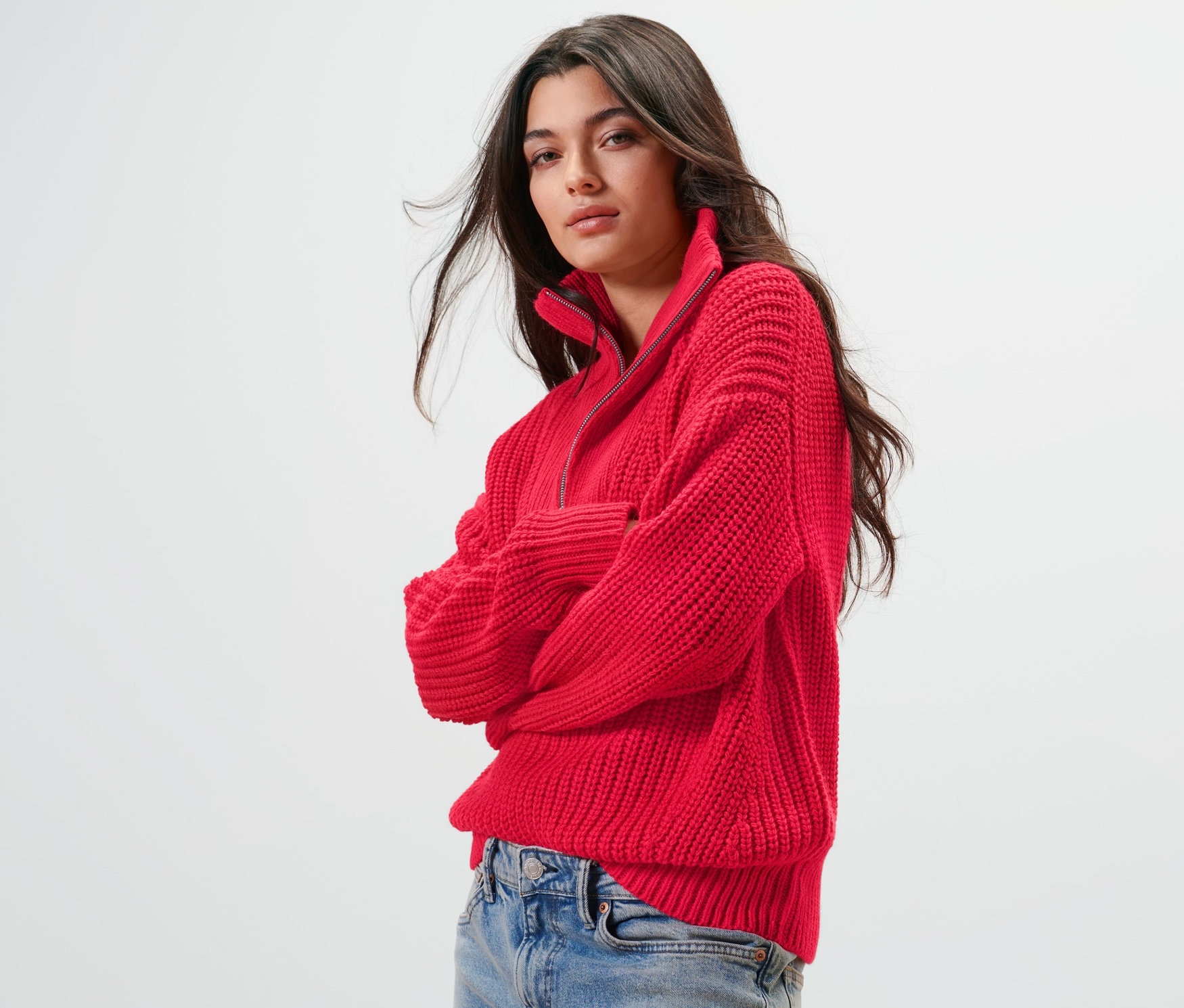Žena má na sobě červený svetr troyer z hrubé pleteniny a džíny.