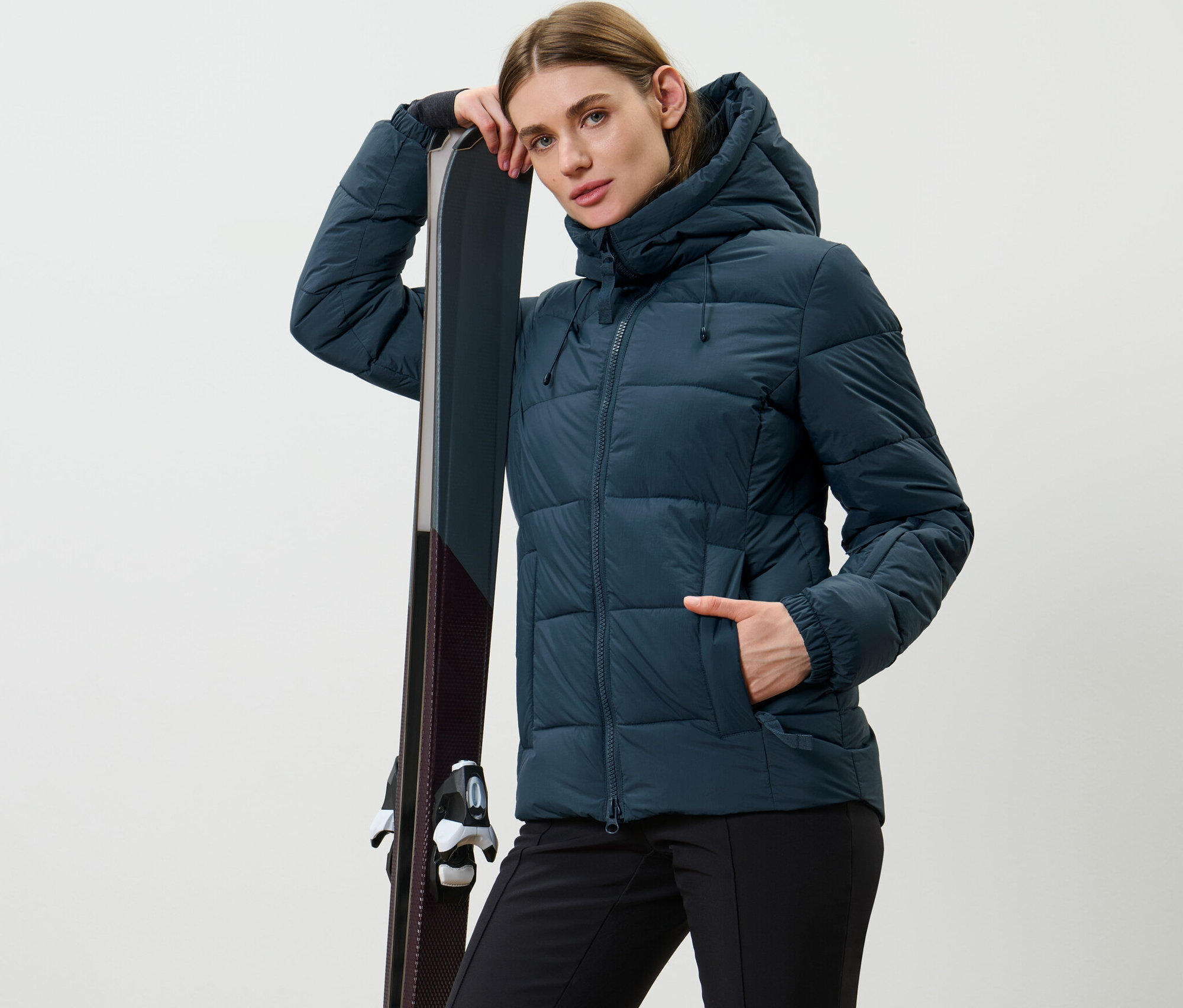 Žena pózuje v tmavě modré prošívané lyžařské bundě a opírá se o lyže.