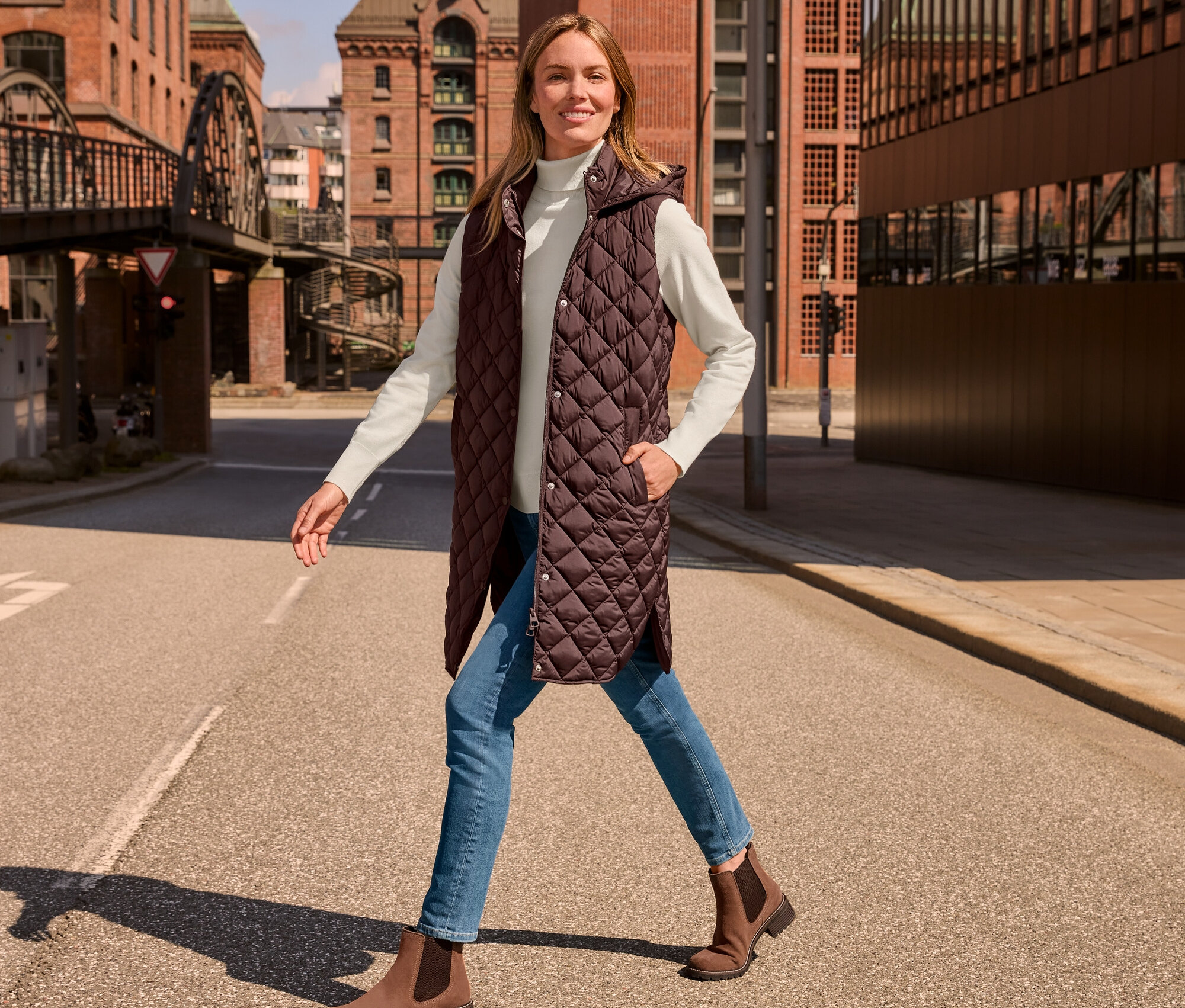 Žena kráčí po ulici a má na sobě dlouhou prošívanou vestu s kapucí, krémově bílý rolák z jemného úpletu, džíny slim – Fit »Emma«, středně modré a semišové boty.