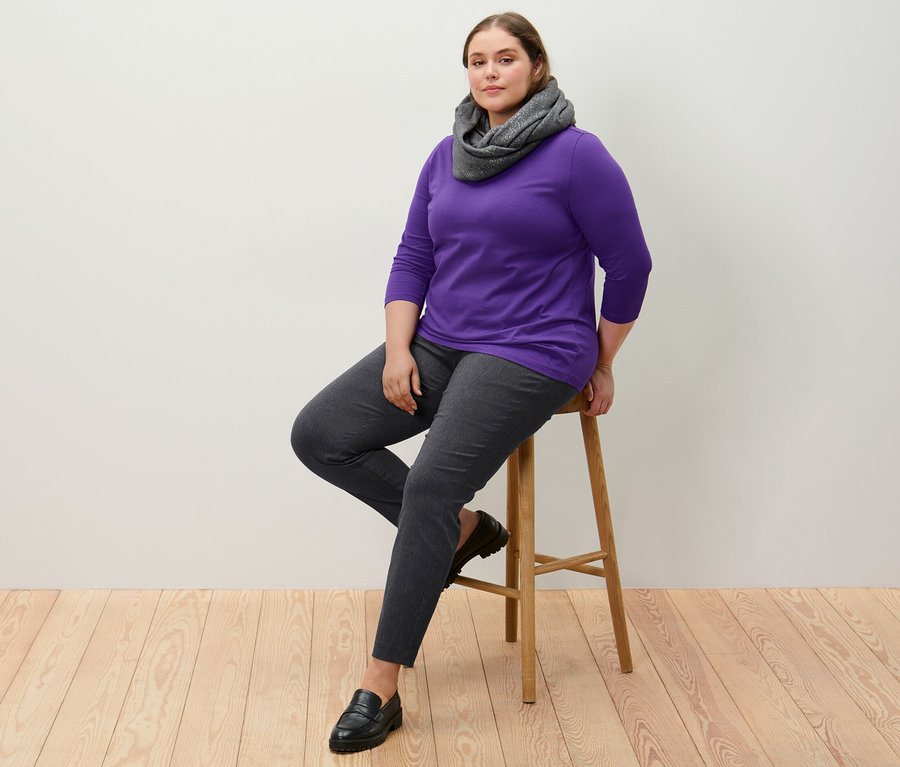 Žena sedí na stoličce v strečových kalhotách a fialovém triku s 3/4 rukávem.