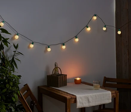 Na zdi visí světelný řetěz s LED. Na stole stojí košík, svíčka a sklenice.