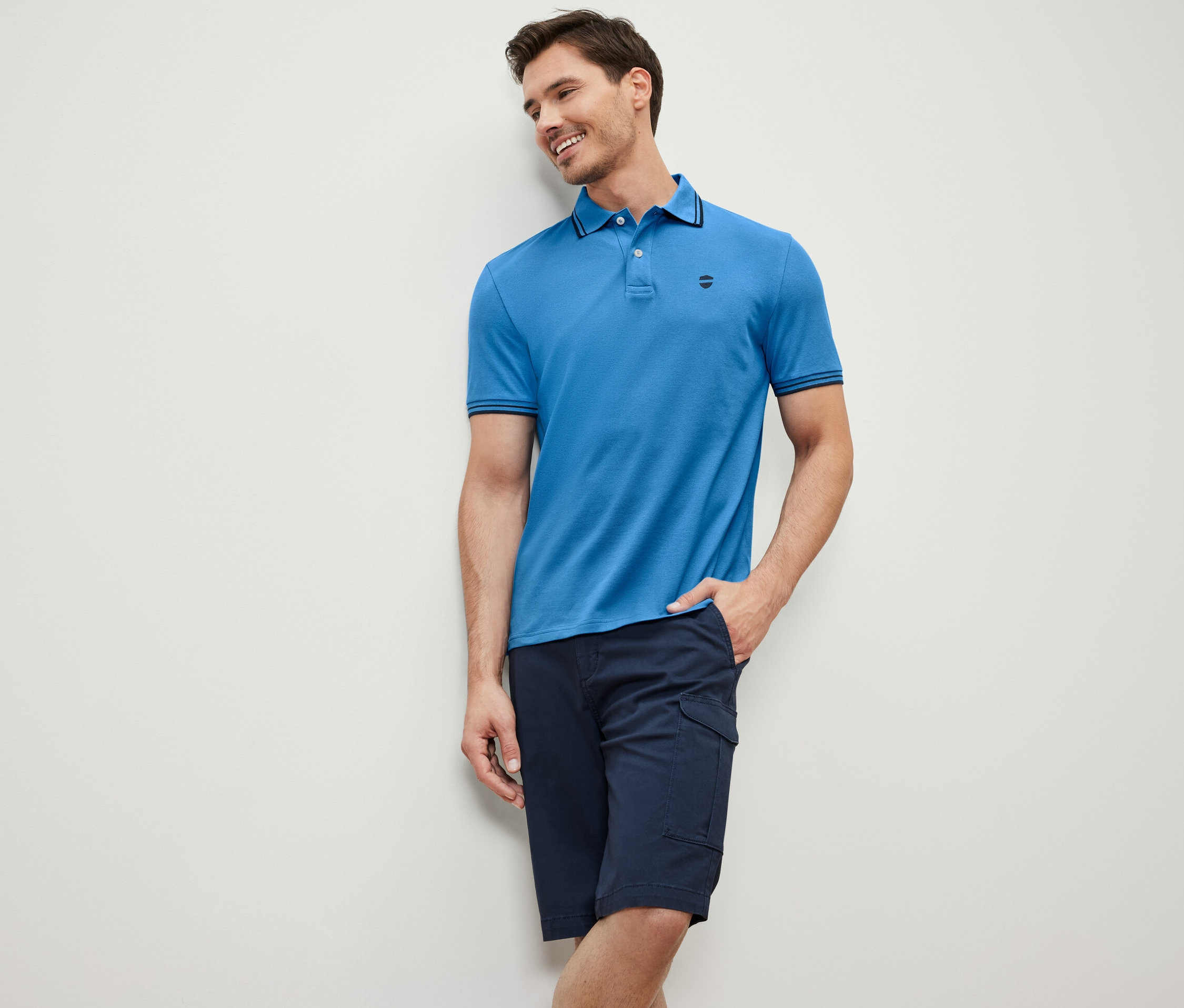 Muž opřený o zeď má na sobě modré polotričko z piké a tmavě modré šortky.