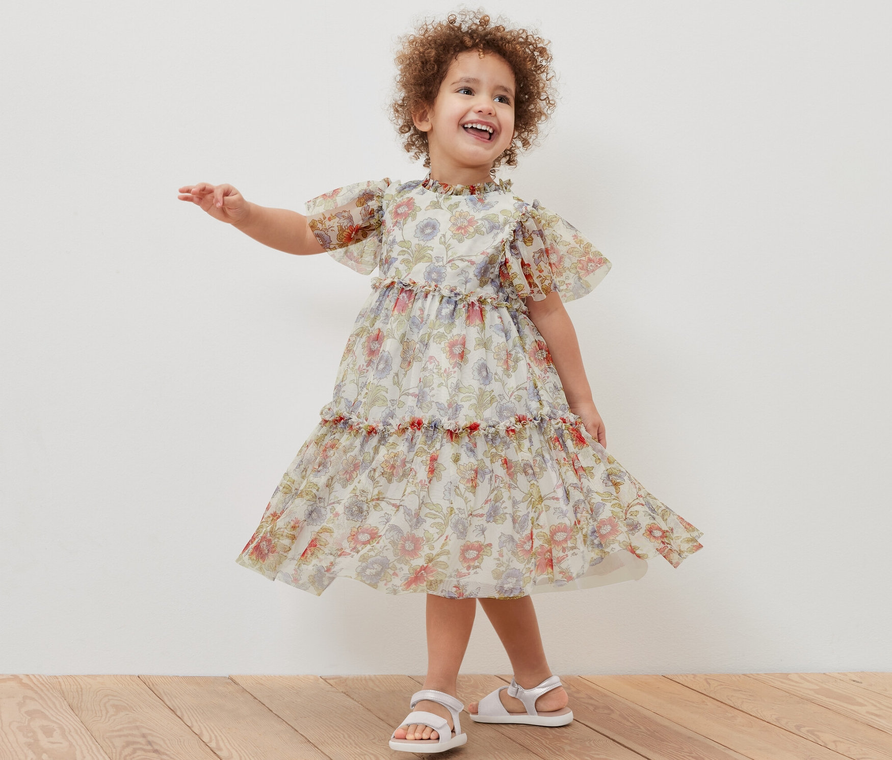 Usmívající se holčička pózuje v tylových šatech s květinovým potiskem a bílých sandálech.