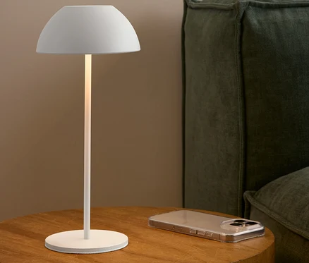 Bílá lampa stojí na dřevěném stolku vedle mobilního telefonu a zelené pohovky.
