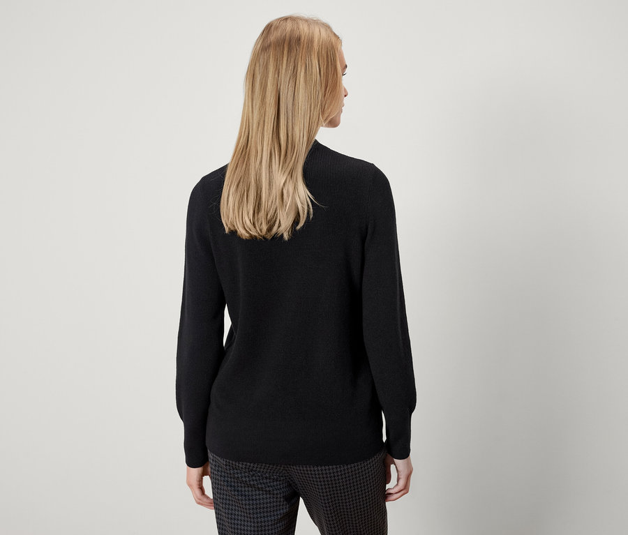 Zezadu je vidět žena s blond vlasy, která má na sobě černý svetr a kalhoty se vzorem.