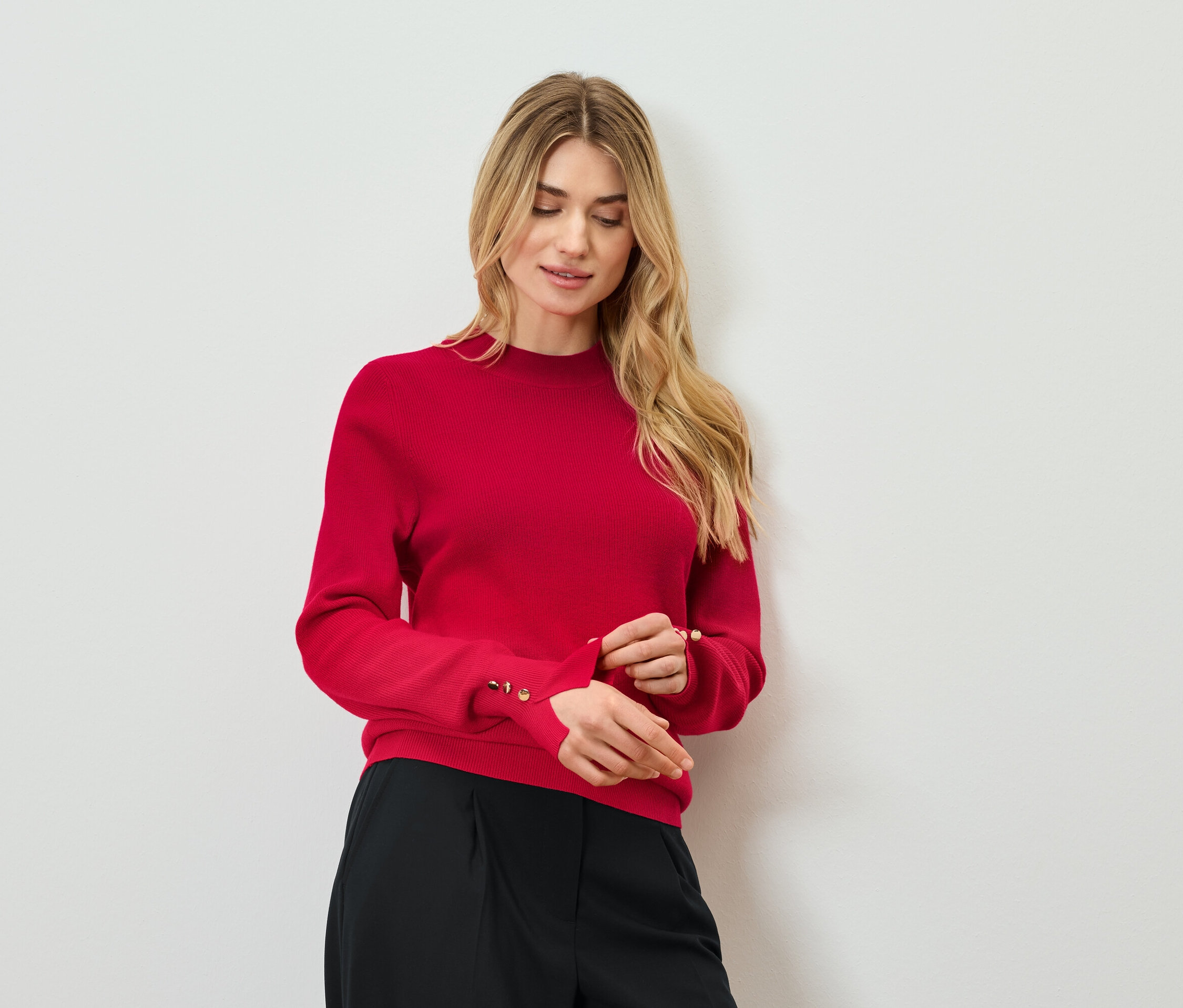 Blondýna si upravuje rukáv červeného svetru s ozdobnými knoflíky. Má na sobě černé kalhoty.