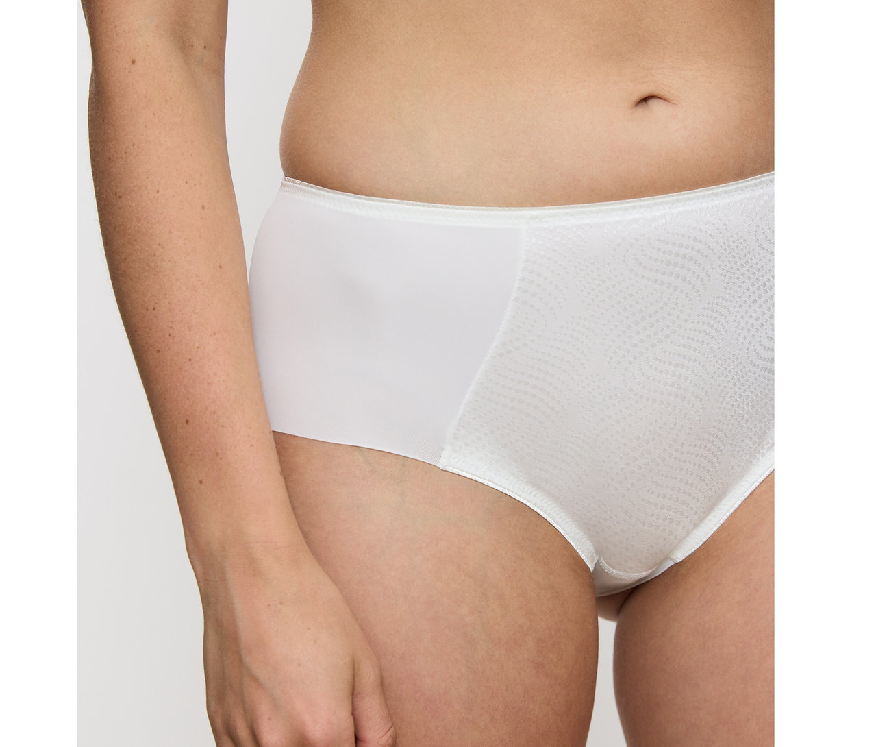 Zblízka osoba v vanilkových zmenšujících bokových kalhotkách Triumph Essential »Minimizer Hipster X«. Kalhotky mají na přední straně texturovaný vlnitý vzor.