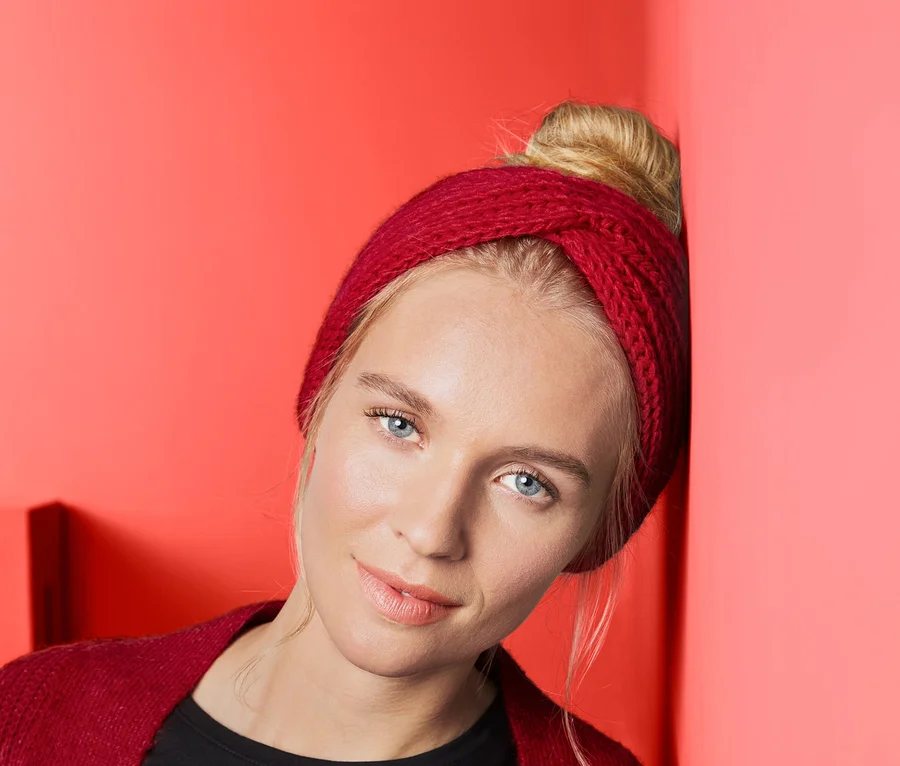 Blondýna opřená o korálově červenou zeď má na sobě červenou čelenku.
