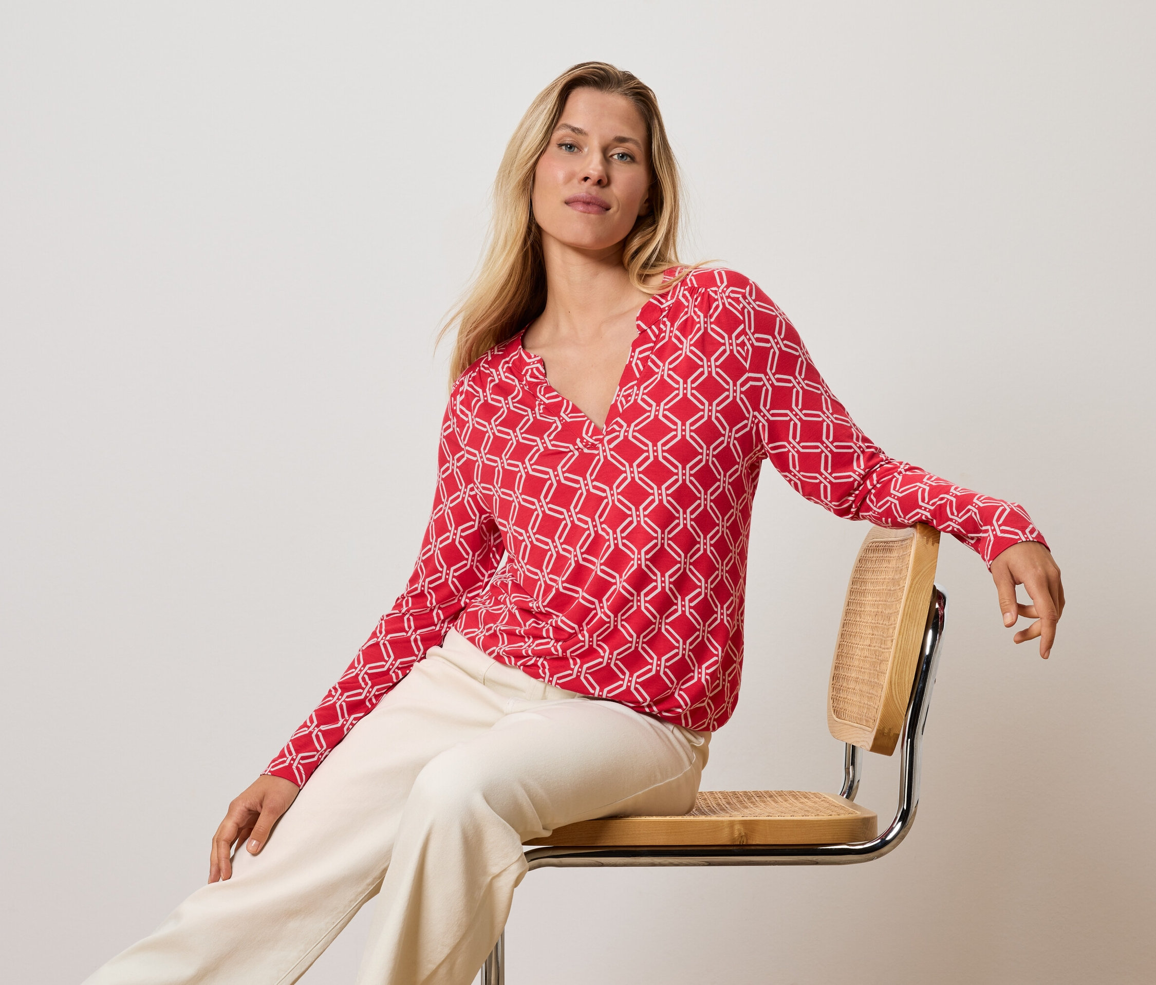 Blondýna sedí na židli, má na sobě červeno-bílý top a krémové kalhoty.