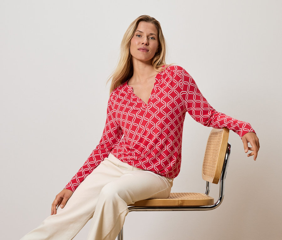 Blondýna sedí na židli, má na sobě červeno-bílý top a krémové kalhoty.