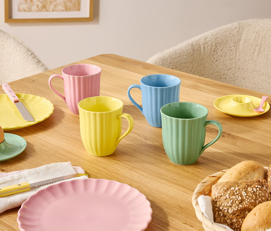 Na dřevěném stole jsou prostřeny talíře a hrnky v pastelových barvách a košík s pečivem.