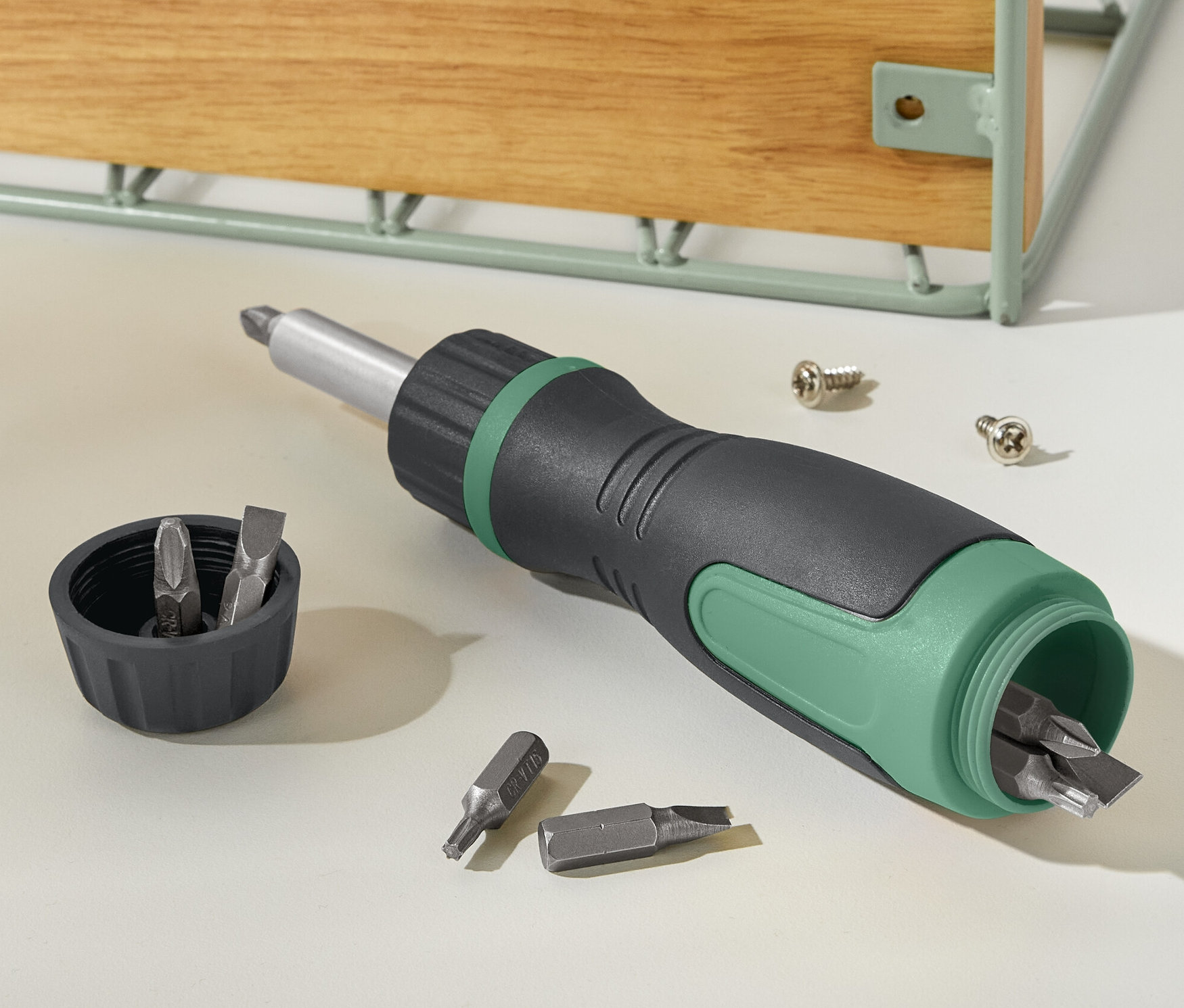 Černo-zelený magnetický ráčnový šroubovák leží vedle víčka s bity a dvou šroubů.