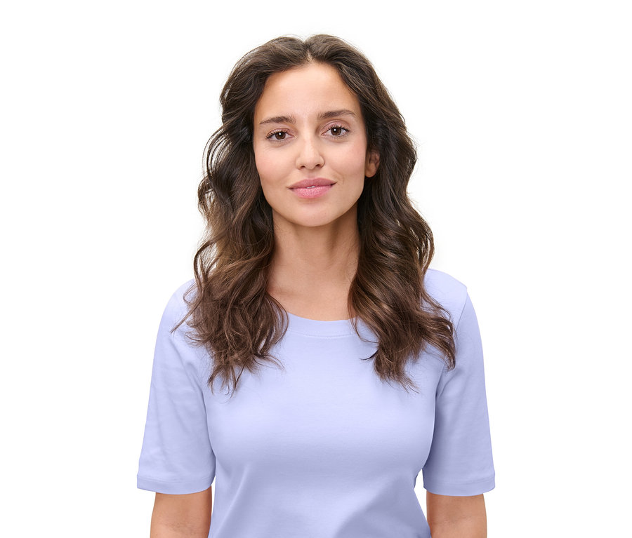 Portrét ženy s hnědými vlnitými vlasy a bleděmodrým tričkem na bílém pozadí.