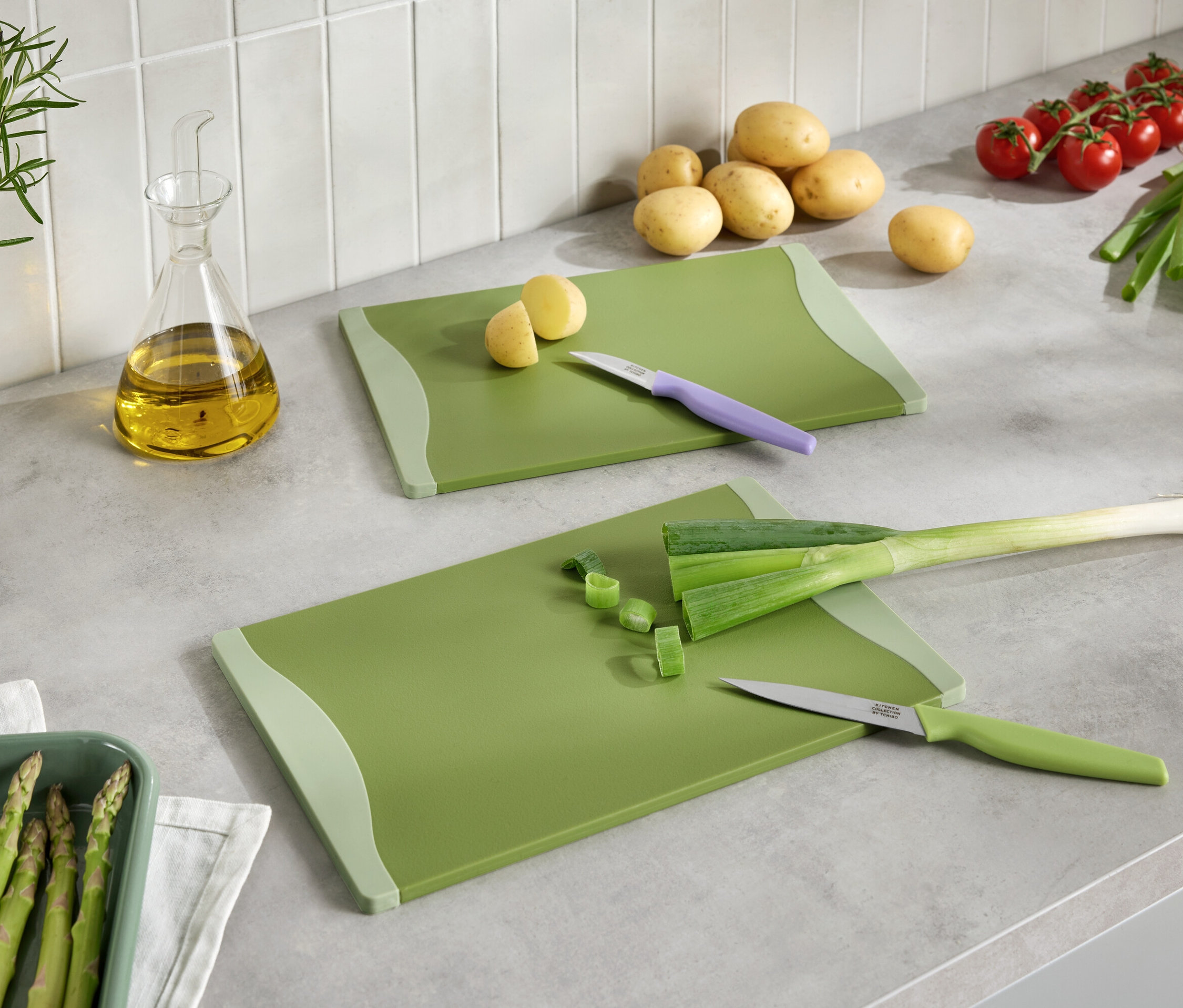 Dvě zelená prkénka s nakrájenou zeleninou, nože, brambory, rajčata, pórek, chřest a olej na kuchyňské lince.